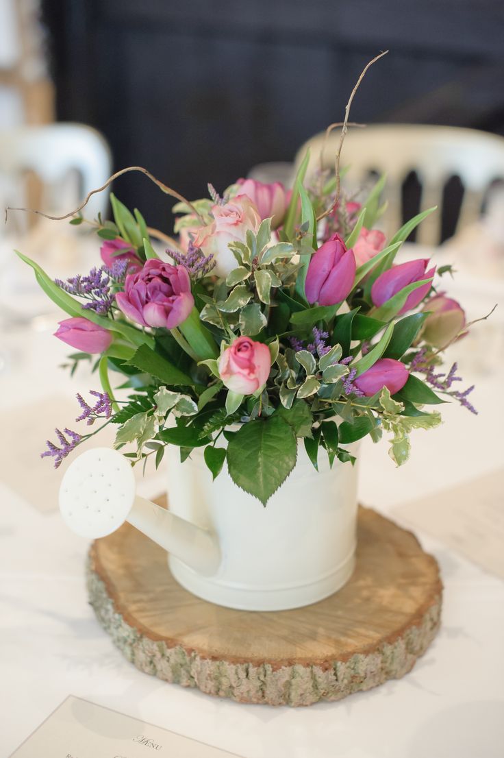 a white coffee cup filled with pink flowers on top of a wooden slice at a table