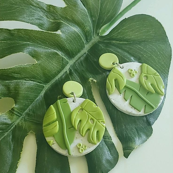 two green and white earrings sitting on top of a large leafy plant next to a pair of scissors