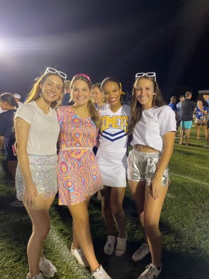 four girls posing for the camera in front of a crowd at a football game, wearing cheerleader outfits