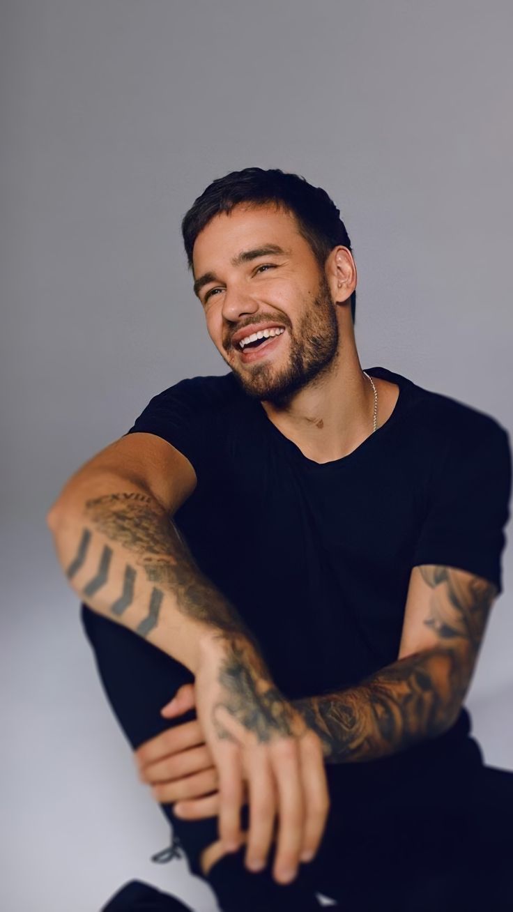 a man sitting on the ground with his arms crossed and tattoos on his chest smiling