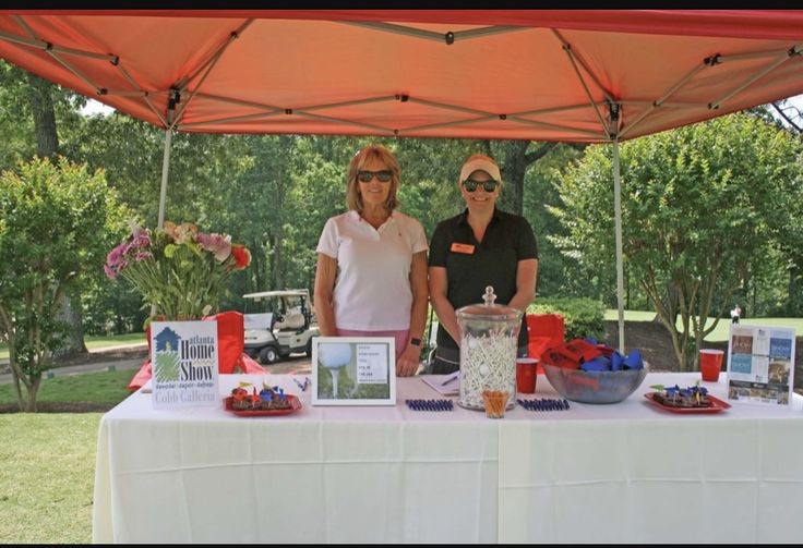 two women standing behind a table with pictures and flowers on it at an outdoor event