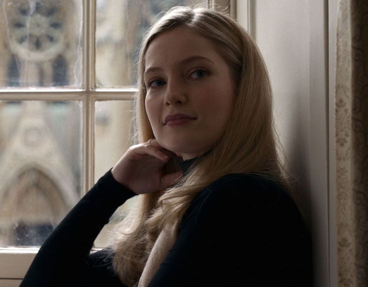 a woman standing in front of a window with her hand on her chin and looking at the camera