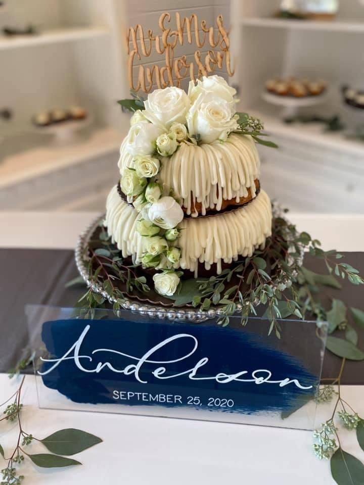 a wedding cake with white flowers and greenery sits on a table in front of an acrylic sign