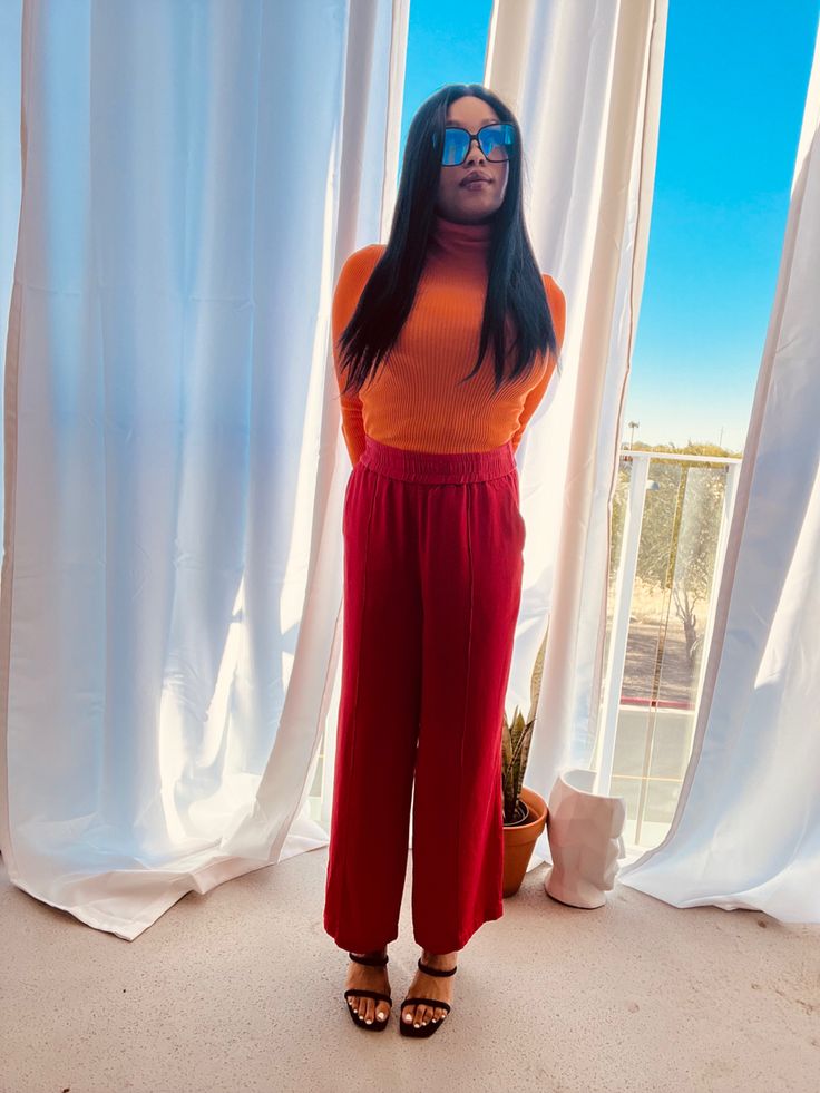 a woman standing in front of a window wearing red pants and an orange sweater with her hands on her hips