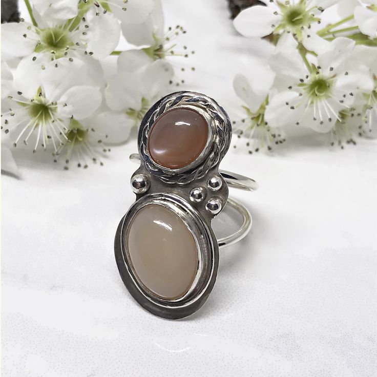 Not only does this ring have a dreamy peach moonstone but it also has an amazing cream moonstone too.  The coloring draws you.  Once you’re there, the pearlescent glow will keep you there.  The peach moonstone is a soft but rich terracotta color.  Equally important is the creamy coloring of the ivory moonstone.  Both stones sit nestled within silver bezel cups.  The peach stone is accented with a hammered silver braid.  A simple wire frame wraps around the ivory stone. Round Cabochon Moonstone Ring, Bohemian Oval Cabochon Moonstone Ring, Unique Open Moonstone Ring With Cabochon Cut, Elegant Untreated Moonstone Ring, Unique Cabochon Chalcedony Ring, Adjustable Cabochon Moonstone Wedding Ring, Unique Handmade Oval Cabochon Moonstone Ring, Handmade Unique Oval Moonstone Ring, Moonstone Rings With Natural Stones