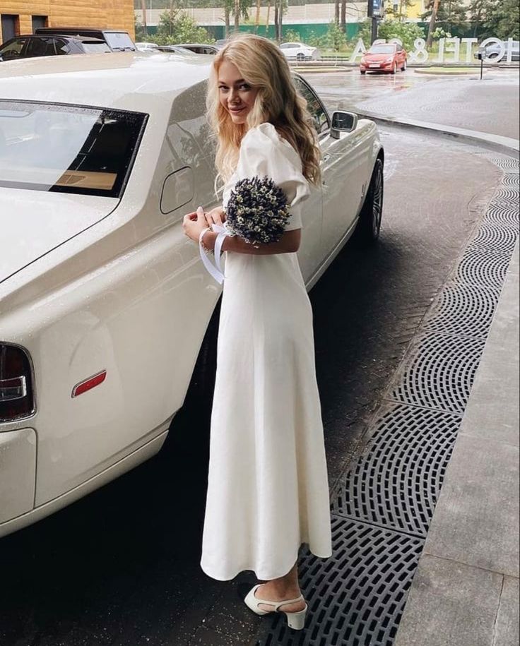 a woman standing next to a white car on the side of the road with her hand in her pocket