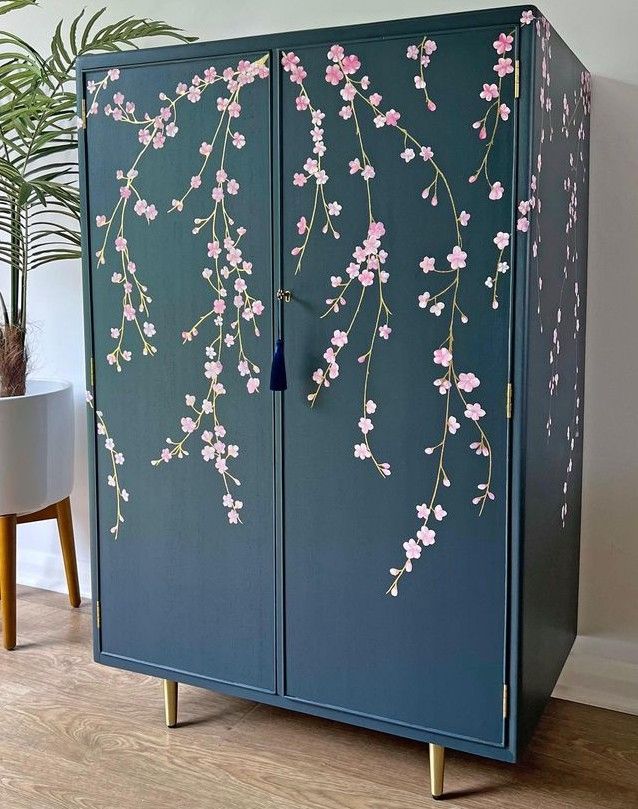 a blue cabinet with pink flowers painted on the side and gold legs, in front of a potted plant