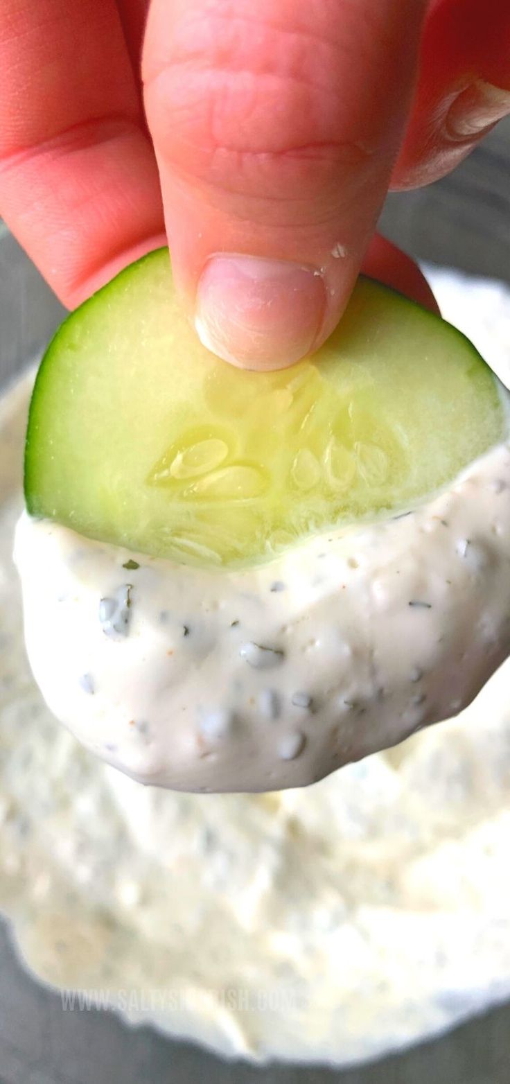 a hand holding a cucumber over a bowl of cream cheese sauce with a pickle in the middle