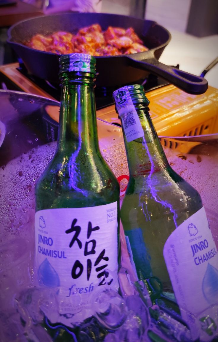 two bottles of beer sitting on top of ice next to a pan filled with food