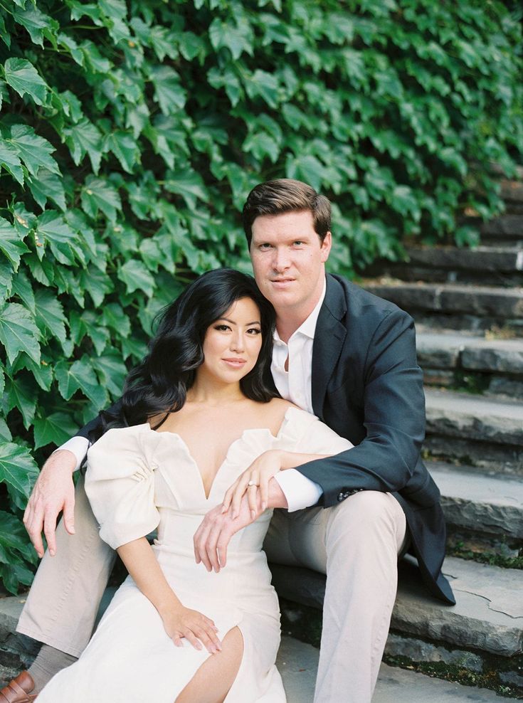 a man and woman are sitting on the steps in front of some bushes with their arms around each other