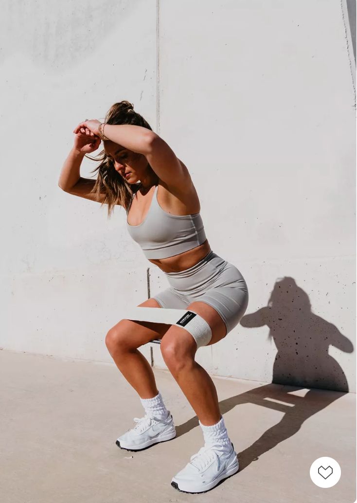 a woman squatting in front of a white wall with her hands behind her head