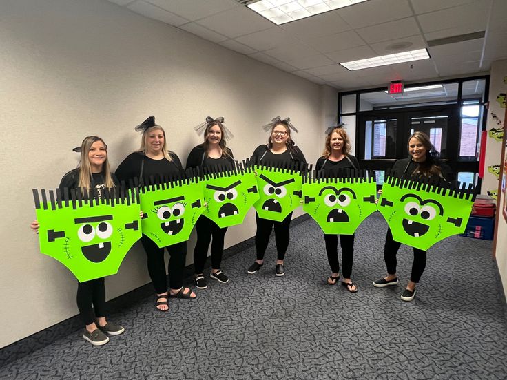 five girls in black shirts are holding up green paper cutouts with faces on them