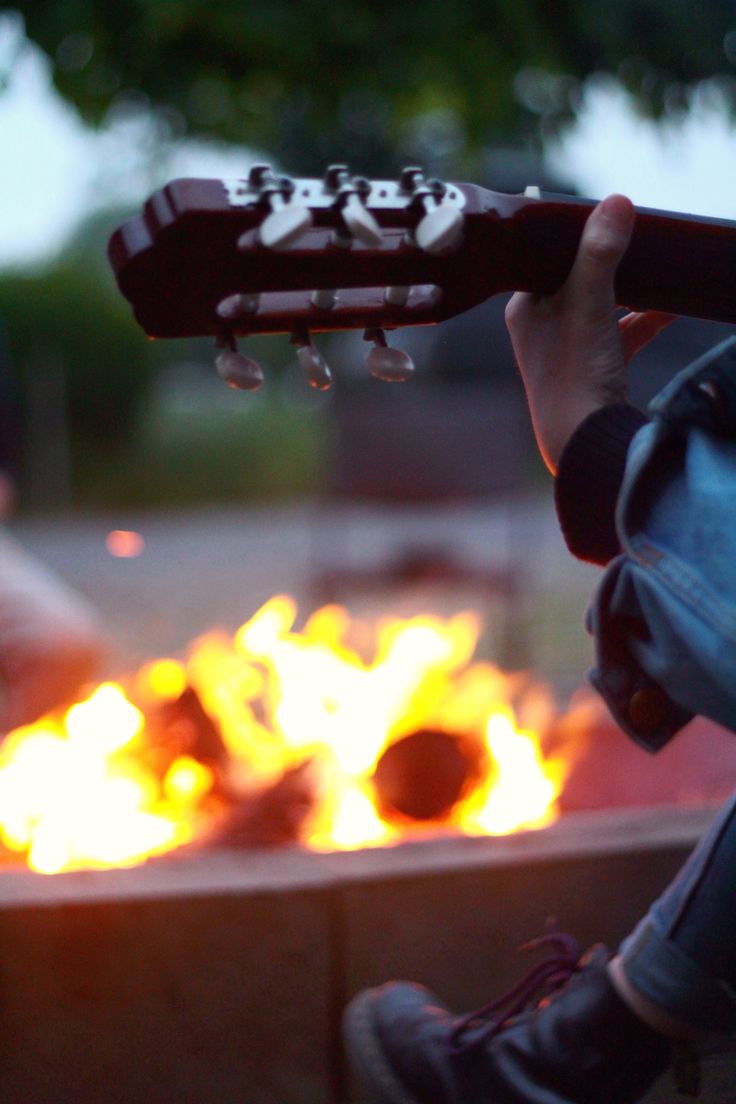 a person playing an acoustic guitar in front of a fire pit with flames behind them