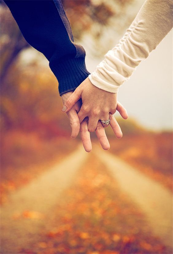 two people holding hands while walking down a dirt road in the fall with leaves on the ground