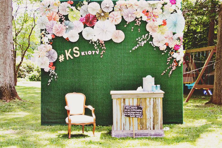 an outdoor photo booth with flowers on the wall and a chair in front of it