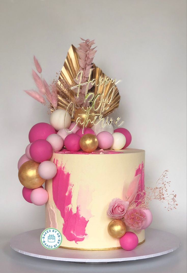 a pink and gold birthday cake decorated with confetti, feathers, and flowers