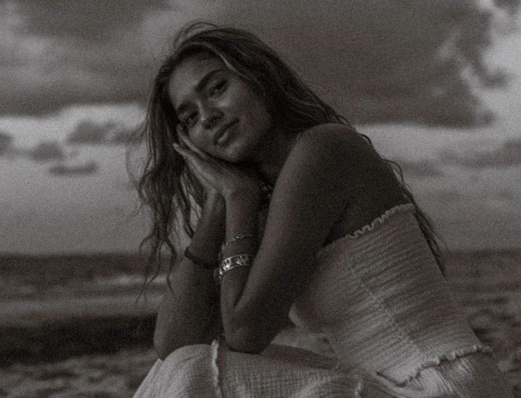 black and white photograph of a woman sitting on the beach with clouds in the background