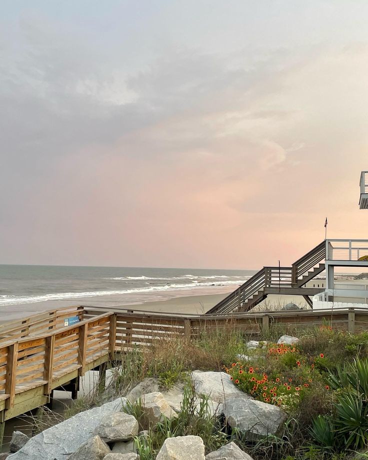 a wooden walkway leading to the beach with flowers growing on it and a life guard tower in the background