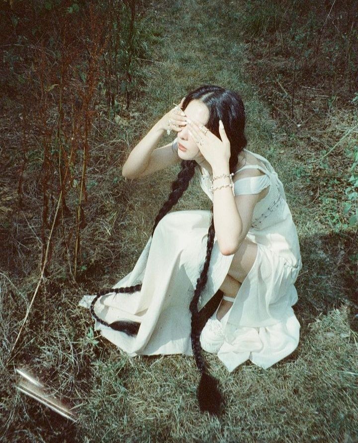 a woman sitting in the grass with her hands on her face and long hair tied to her head