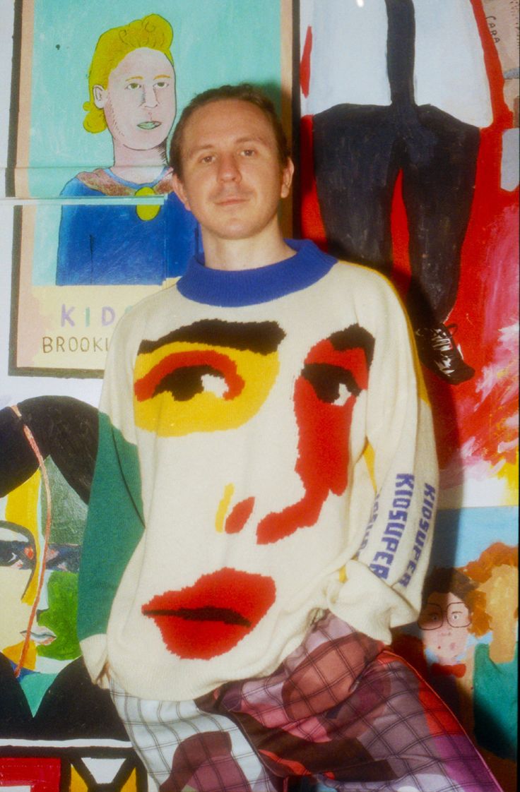 a man standing in front of a wall with paintings on it and wearing a sweater