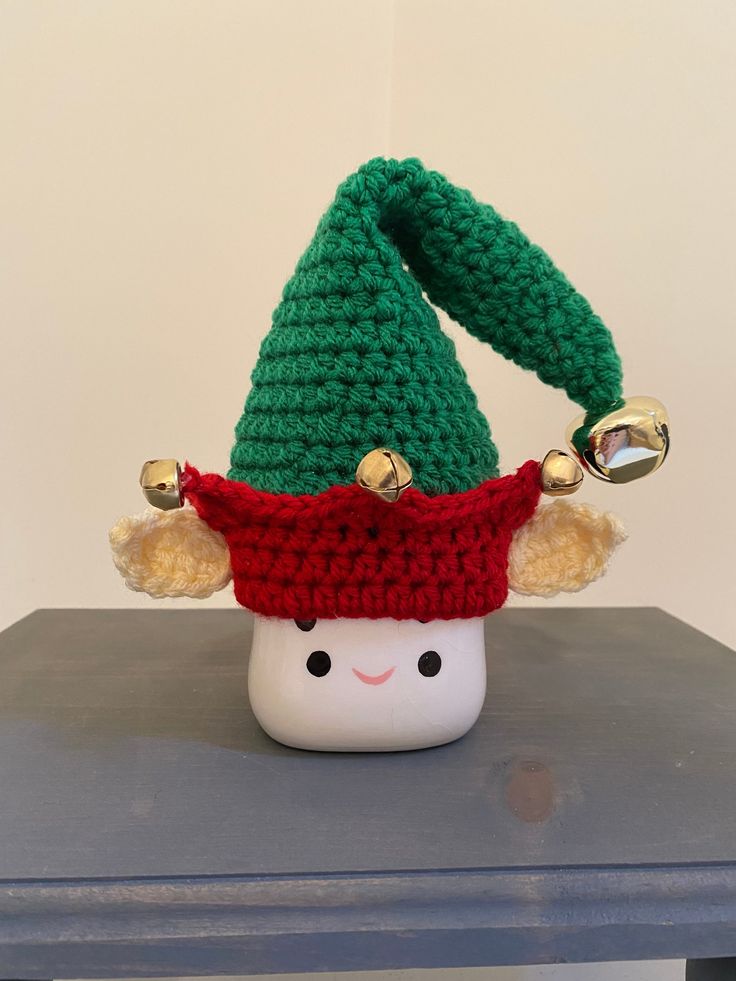 a crocheted christmas hat with bells on top of a small white cup sitting on a table