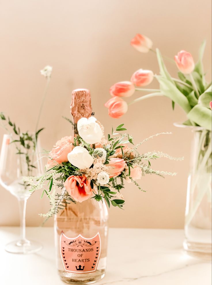 an arrangement of flowers in a bottle on a table with two wine glasses and a vase