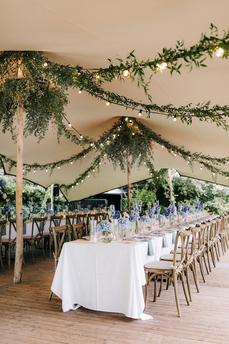 an outdoor tent with tables and chairs set up for a formal dinner or party, decorated with greenery hanging from the ceiling