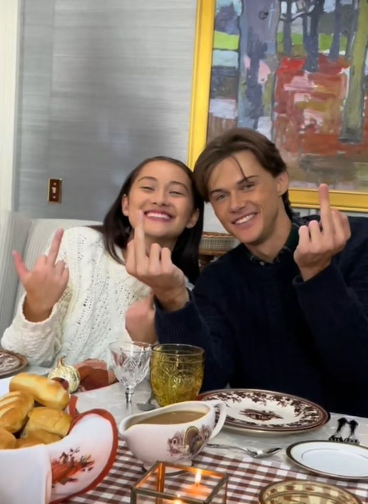 two people sitting at a table with food and drinks in front of them, making the peace sign