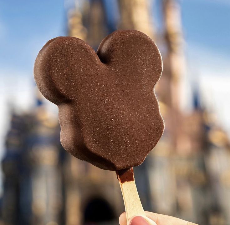 a hand holding a chocolate mickey mouse lollipop in front of a castle like building