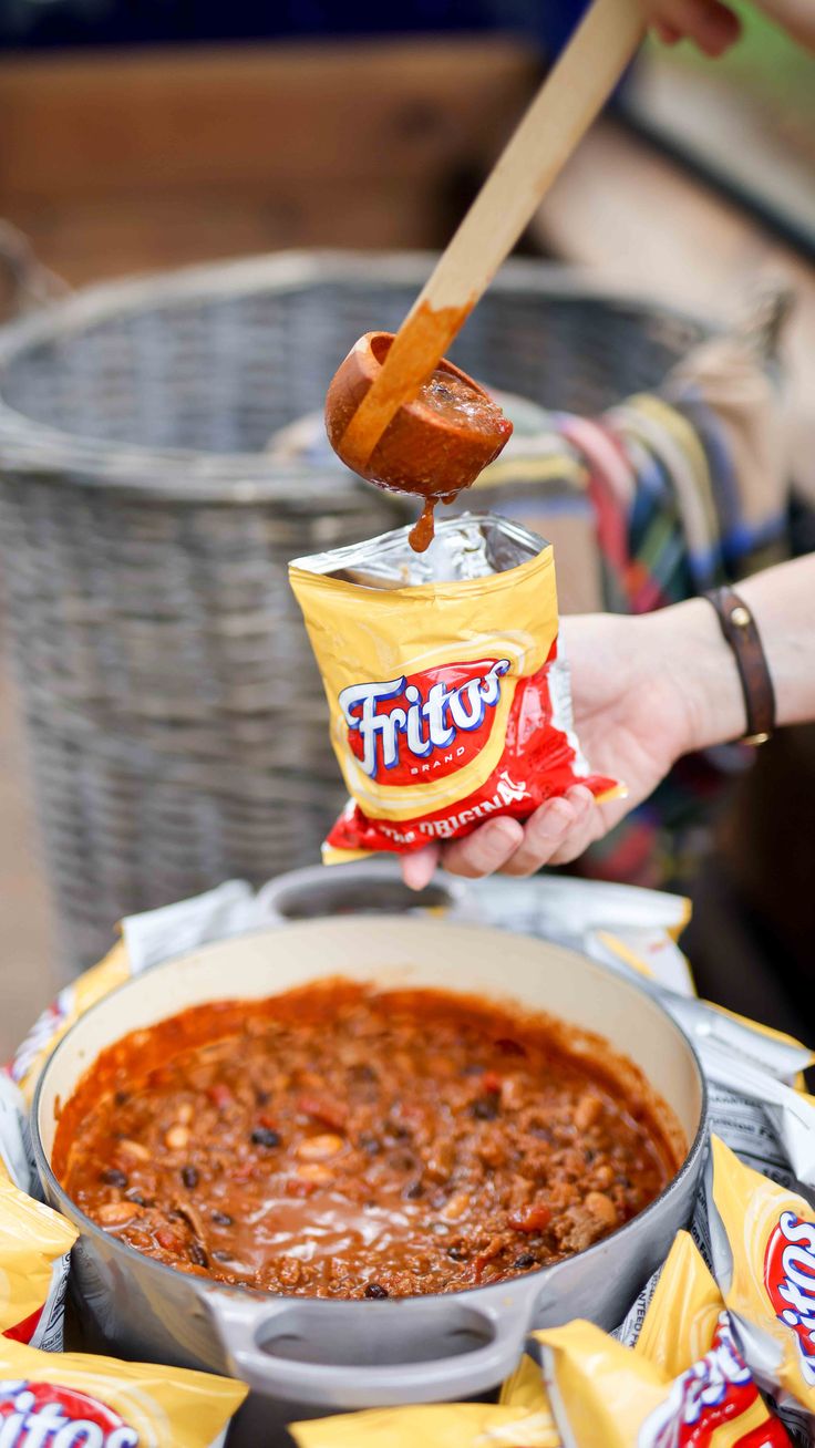 a person holding a bag of frito's chili next to a pot of chili