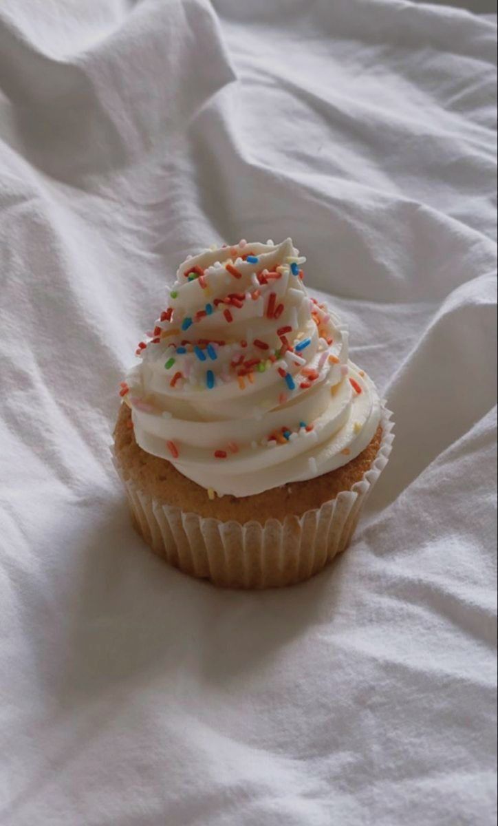 a cupcake with white frosting and sprinkles sitting on a bed