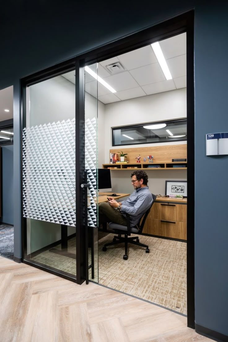 a man sitting at a desk in an office cubicle looking out the glass door