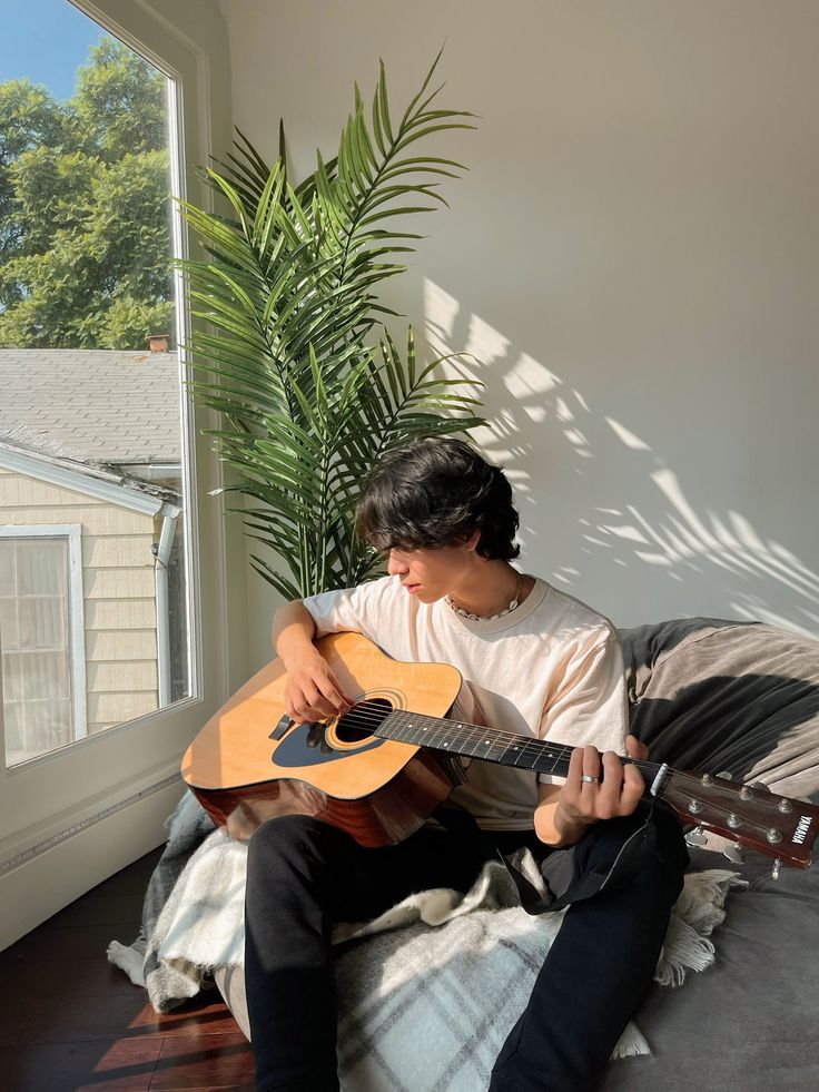 a young man sitting on top of a bed holding an acoustic guitar next to a window