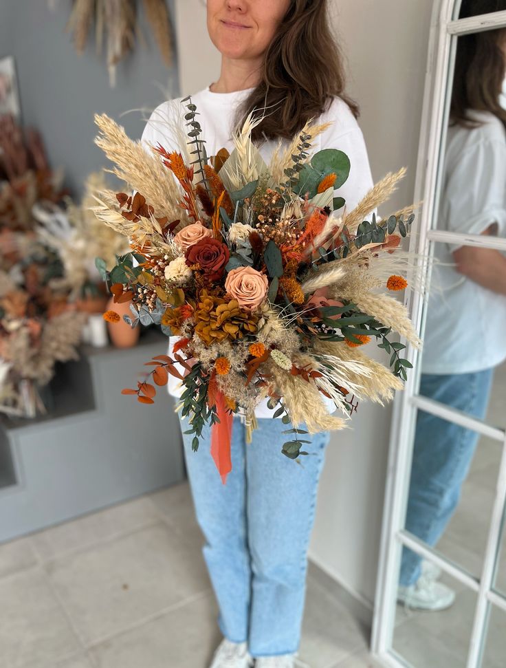 a woman standing in front of a mirror holding a bouquet of dried flowers and foliage