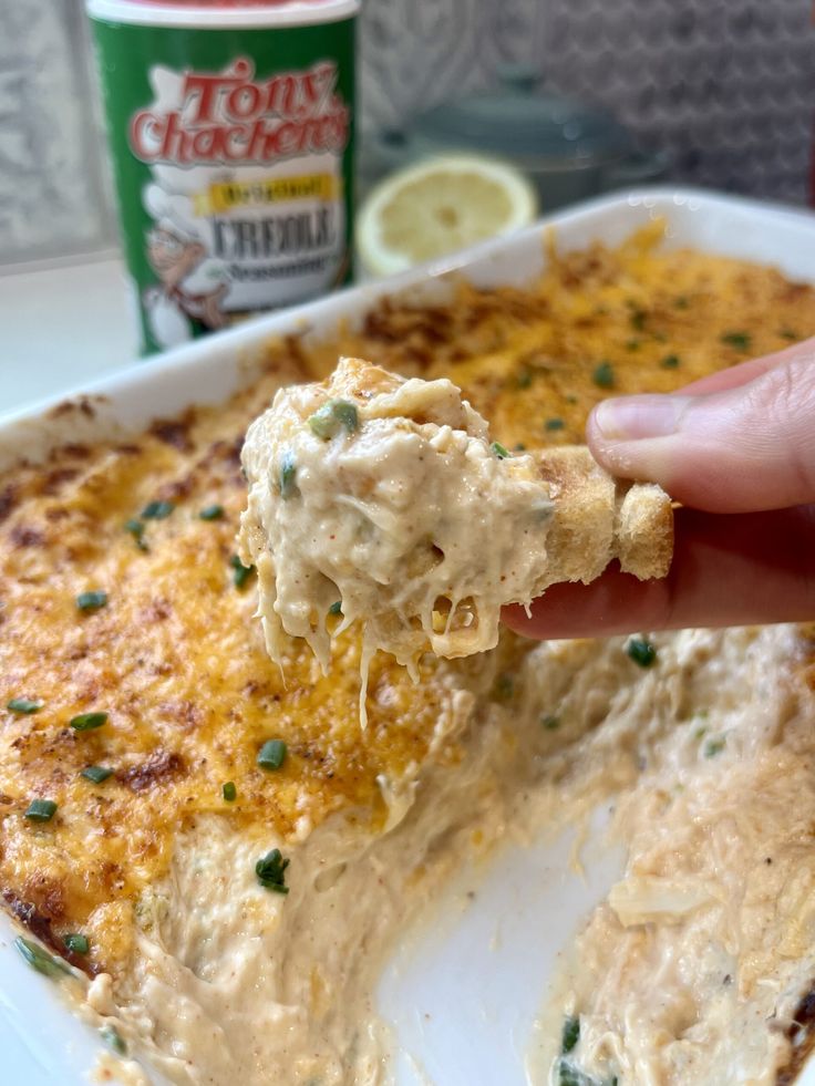 a hand holding a piece of food over a casserole dish with cheese and green onions