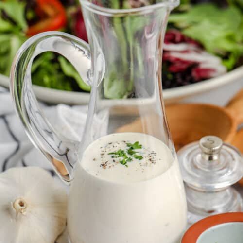 a salad with dressing in a glass pitcher next to a bowl of greens and garlic