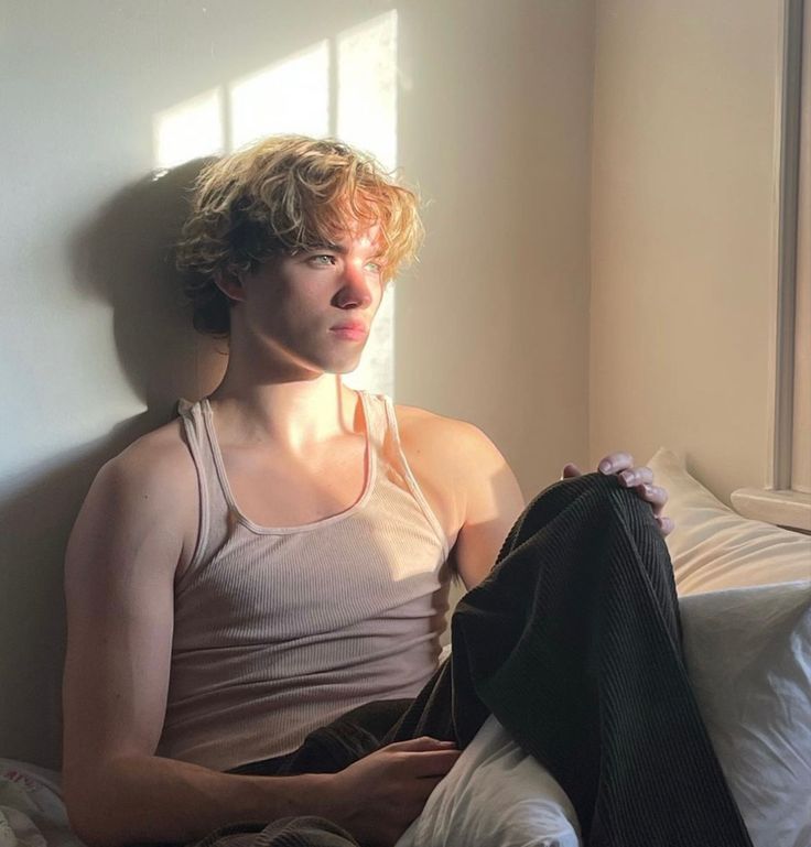 a young man sitting on top of a bed next to a window with sunlight streaming through it