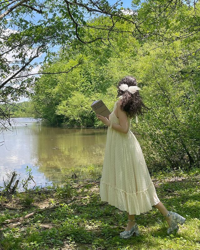 a woman in a yellow dress reading a book by the water with trees around her