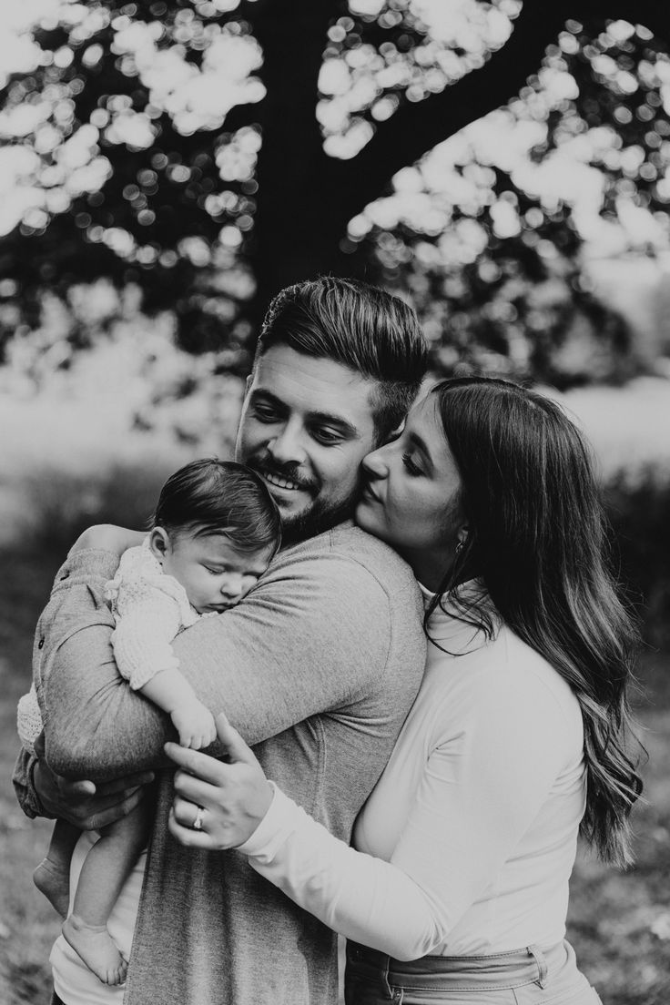 a man and woman holding a baby while standing next to each other in front of a tree