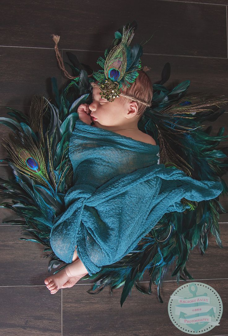 a baby wrapped in a peacock costume laying on top of a wooden floor with feathers around it