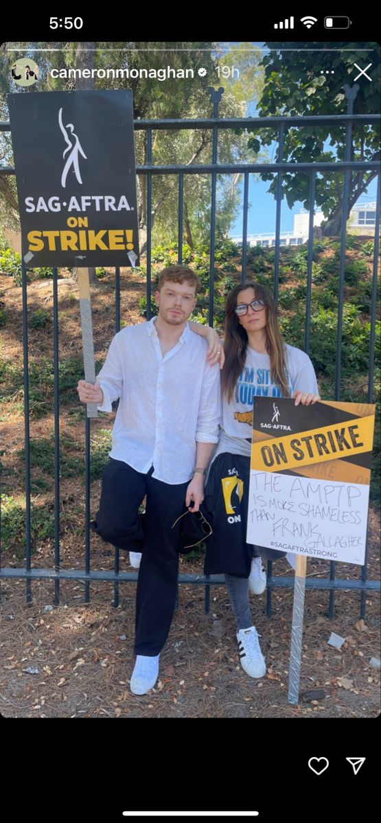 two people standing next to each other in front of a fence with signs on it