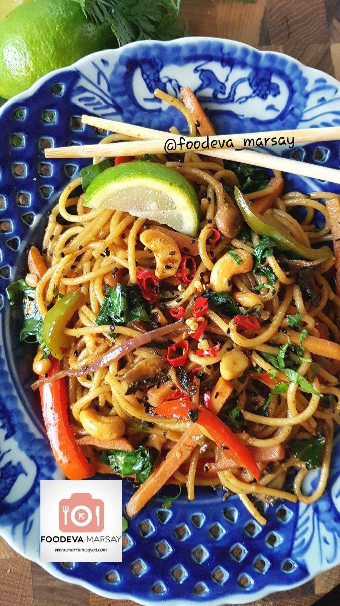 a blue and white plate topped with noodles, vegetables and chopsticks next to an avocado