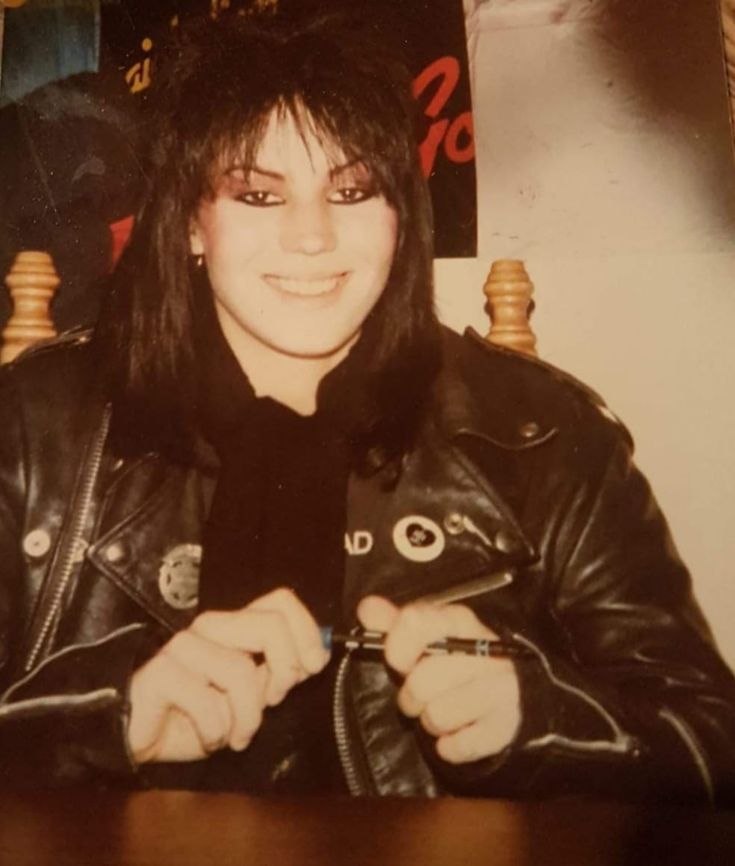 a woman sitting at a table with a leather jacket on and her hand in her pocket