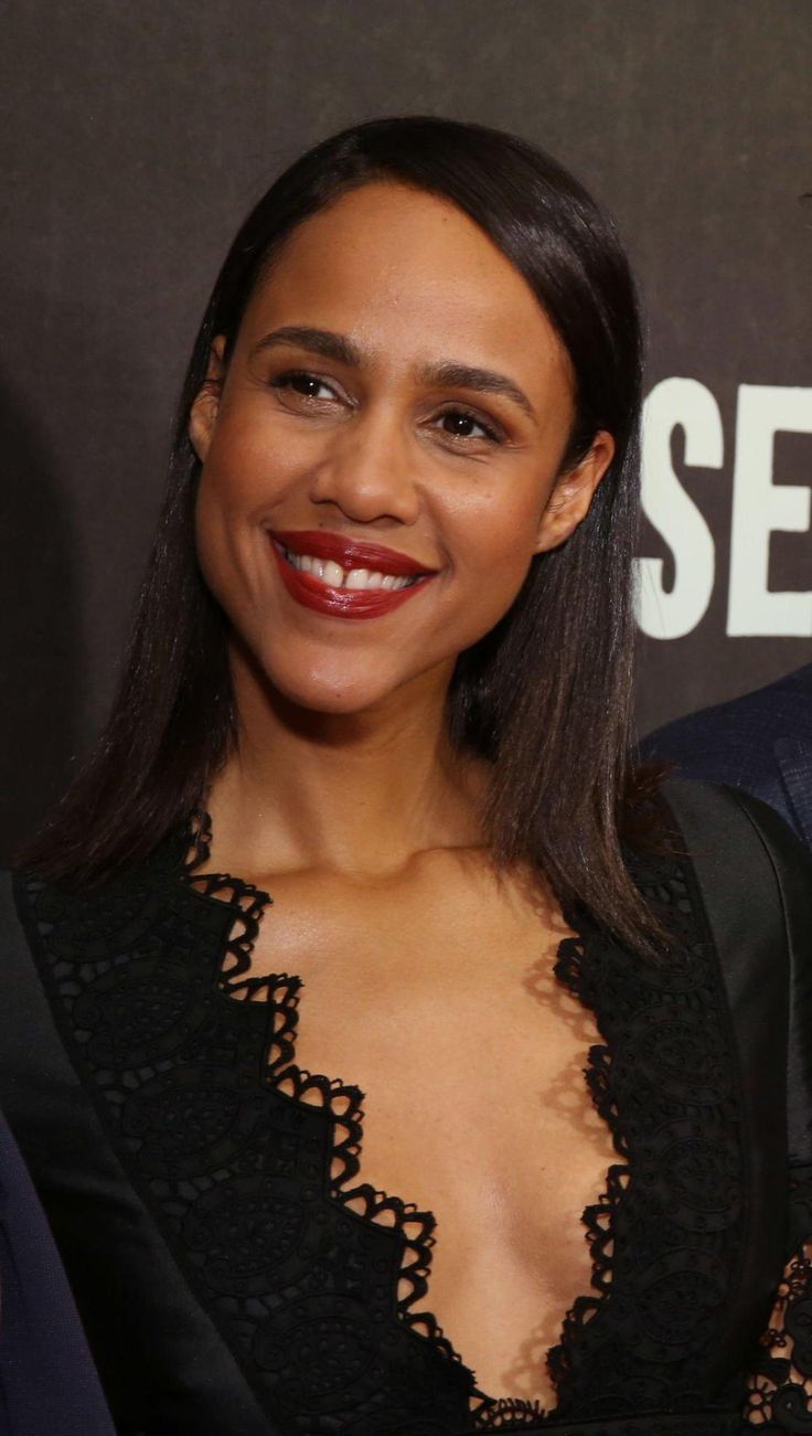 a smiling woman in a black dress and red lipstick poses for the camera with another man behind her