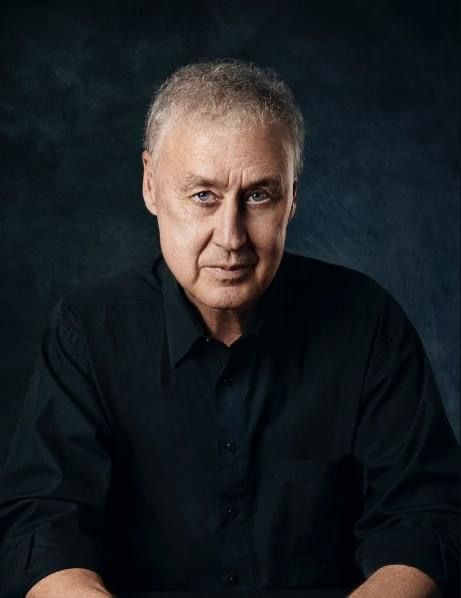 an older man sitting at a table with his hands on his knees and looking into the camera