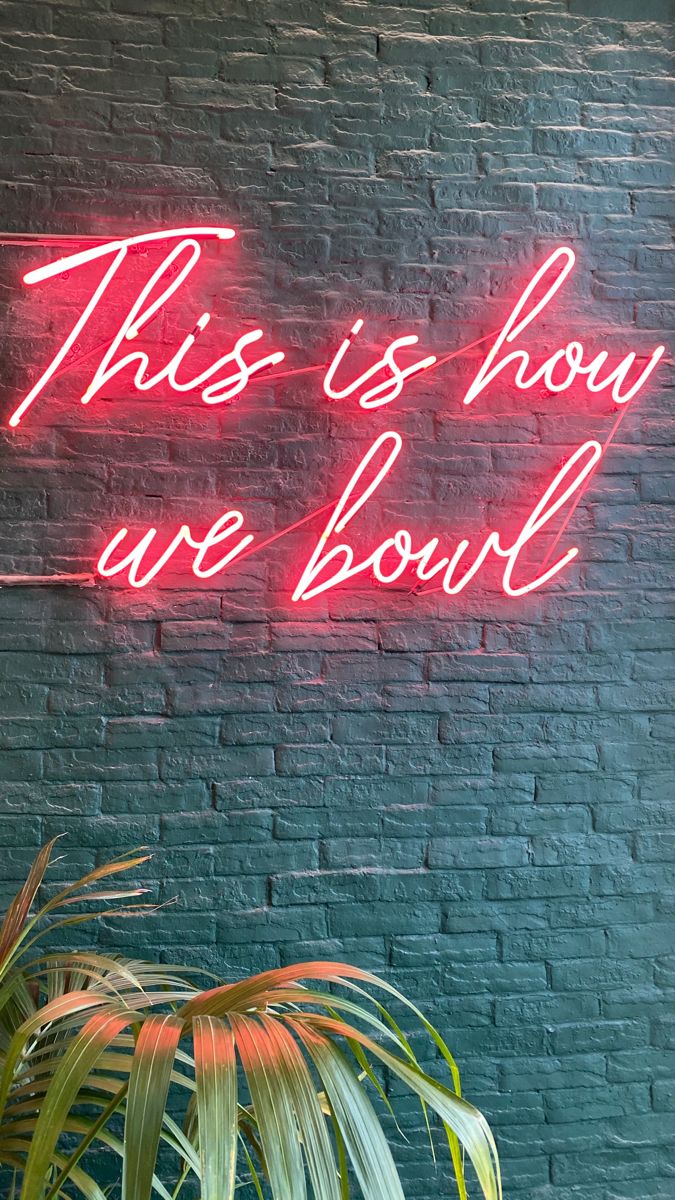 a neon sign that says this is how we bowl on a brick wall next to a potted plant