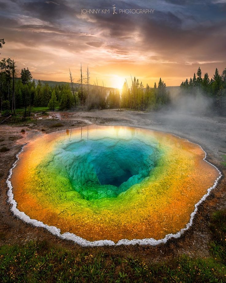 a rainbow colored pool in the middle of a forest with trees and clouds behind it