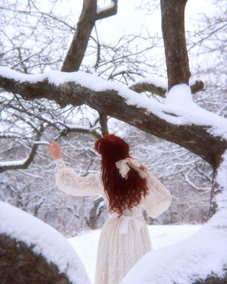 a woman with red hair is standing in the snow and looking up at some trees