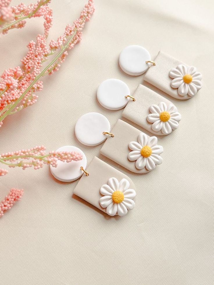 four white and yellow flower shaped earrings sitting on top of a table next to pink flowers