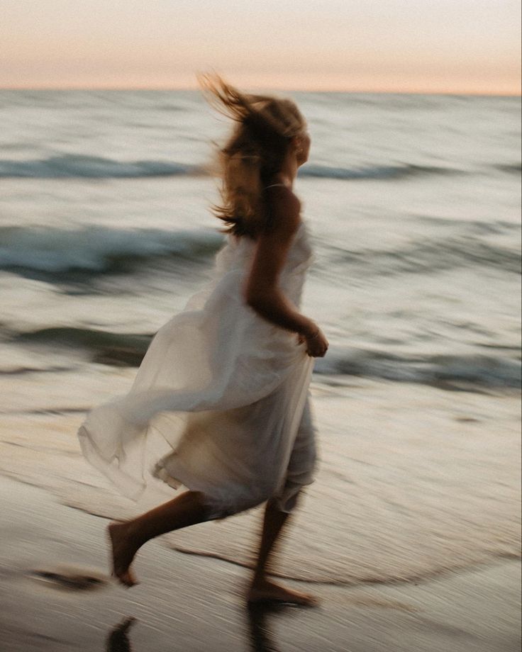 a woman in white dress running on beach next to the ocean with waves coming in